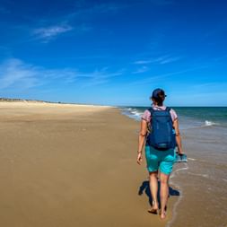 Wanderin mit blauem Rucksack läuft barfuß am Sandstrand entlang mit türkisfarbenen Meereswellen unter blauem Himmel am Portugiesischen Jakobsweg.
