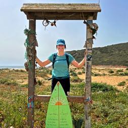 Lächelnde Wanderin in türkisfarbenem Shirt und Kappe steht in Holzrahmen mit grünem Surfboard am Praia do Barranco, Algarve, Küstenhügel dahinter.