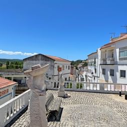 Cobblestone square in Aljezur with white balustrade, stone monument, and traditional Portuguese buildings with terracotta roofs under blue sky.