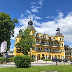 Yellow baroque castle hotel with ornate towers and balconies in Velden, surrounded by green trees and lawn under a blue cloudy sky.