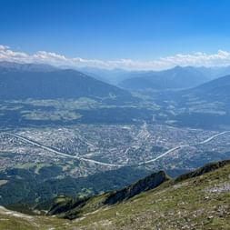 Luftaufnahme der Stadt Innsbruck im Inntal, umgeben von Bergketten. Grüne Almwiesen im Vordergrund, blauer Himmel mit Wolken darüber.