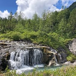 Wasserfall, der über Felsvorsprünge in ein türkisfarbenes Becken fließt, umgeben von grünem Wald und Bergen unter blauem Himmel mit weißen Wolken.