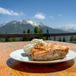 Stück Apfelstrudel mit Schlagsahne auf weißem Teller auf einer Bergterrasse, mit schneebedeckten Alpengipfeln im Hintergrund.