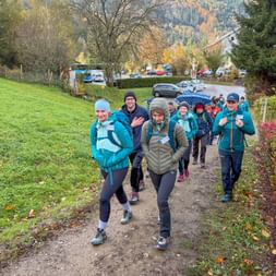 Group of hikers in autumn clothing walking on a path through a village with green meadows and mountains in the background.