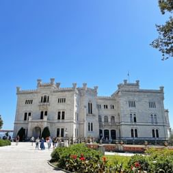 Weißes neugotisches Schloss Miramare mit Zinntürmen und Bogenfenstern, umgeben von gepflegten Gärten mit roten Blumen und Besuchern.