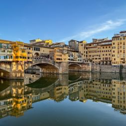 Ponte Vecchio mit historischen Gebäuden über dem Arno in Florenz. Die Steinbrücke und bunten Gebäude spiegeln sich im ruhigen Wasser.