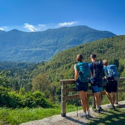 Three hikers with backpacks standing at wooden viewpoint overlooking forested mountains in the Julian Alps near Bovec, Soča Valley.