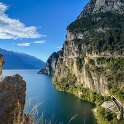 Panoramic view of Strada del Ponale path carved into steep cliffs above Lake Garda, with dramatic rock formations and blue water below.