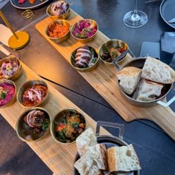 Two wooden serving boards with various colorful mezze dishes in metal bowls, flatbread, and wine glasses on a dark restaurant table.