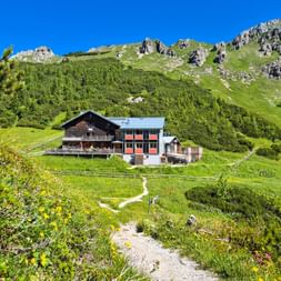 Carl von Stahl Haus mountain hut nestled in green Alpine meadows with rocky peaks and forest. A hiking trail leads to the building.