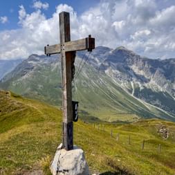 Hölzernes Gipfelkreuz am Sandjöchl mit Panoramablick auf Alpengipfel und grüne Täler unter blauem Himmel mit weißen Wolken.