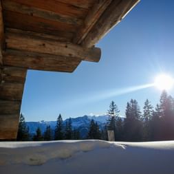 Holz-Berghütte in Blockbauweise mit Blick auf verschneite Landschaft. Nadelbäume und Berggipfel unter blauem Himmel mit Sonne sichtbar.