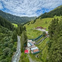 Aerial view of Baumzipfelweg adventure park in Saalbach-Hinterglemm with long slides, a stream, and alpine chalets in a green mountain valley.