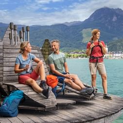 Three hikers resting on a wooden pier at Wolfgangsee in Salzkammergut. Mountains and turquoise lake water visible in the background.