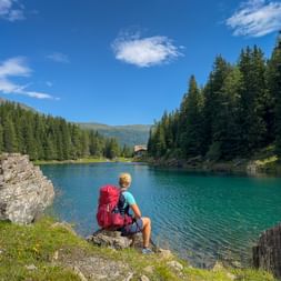 Hiker with red backpack sitting by turquoise Obernberger See, surrounded by forested mountains and blue sky with a small building visible.
