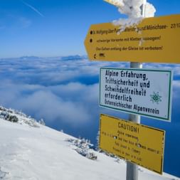 Wegweiser am verschneiten Schafberg mit Wolken darunter. Gelber Richtungsweiser und weiß-grünes Alpenverein-Sicherheitsschild vor blauem Himmel.