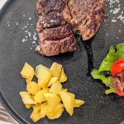 Sliced grilled steak with coarse salt on a dark plate, served with roasted potatoes and garnished with lettuce and red pepper.