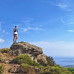 Woman stands in sportswear on a rock high above the sea, blue sky, sunshine