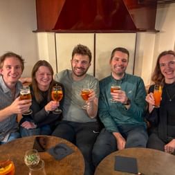 Five colleagues sitting together on a boat, raising glasses with drinks in a toast. They're in an elegant interior with paintings on the walls.