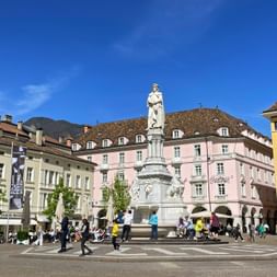 Walther Square in Bozen with white statue monument in center, surrounded by colorful historic buildings with arcades and people walking.
