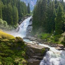 Kraftvoller Wasserfall über moosbewachsene Felsen im Nationalpark Hohe Tauern, umgeben von dichtem Nadelwald und Bergen.