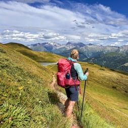 Hiker with red backpack and trekking poles on grassy mountain trail near Obernberg, with alpine peaks and blue sky in background.