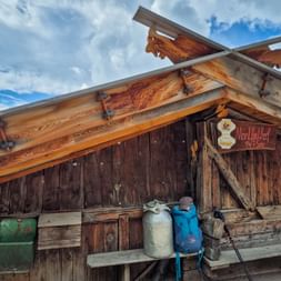 Blue hiking backpack resting on wooden bench outside Montferthof mountain hut with traditional wooden construction and decorative roof beams.