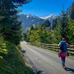 Hiker with backpack walking on gravel path between Fieberbrunn and Saalbach. Snow-capped mountains and forests under blue sky.