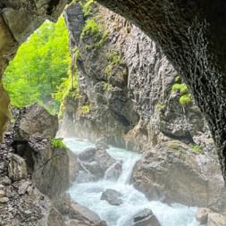 Blick aus einer Höhlenöffnung in der Partnachklamm mit türkisfarbenem Wasser über Felsen und grüner Vegetation draußen sichtbar.