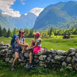 Two hikers sitting on a stone wall in Trenta, Soča Valley. Green meadows and forested mountains of the Julian Alps rise in the background.