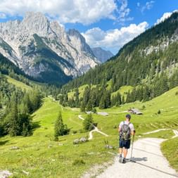 Hiker with backpack walking on paved path through green alpine valley in Weissbach, surrounded by dramatic limestone peaks and forests.