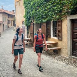 Two female hikers with backpacks walking through a cobblestone alley in Monforte, Piedmont. Ivy-covered buildings and traditional houses line the street.