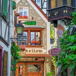 Narrow alley in Rüdesheim's Drosselgasse with colorful half-timbered buildings, decorated facades, cafe signs, and flower boxes on windows.