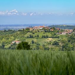 View of Baudra village on a hilltop in Piedmont with green fields, vineyards, and snow-capped Alps in the distance under blue sky.