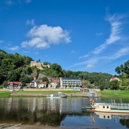 Fährboote an der Fährstelle Rathen an der Elbe mit Dorfhäusern und bewaldeten Hügeln. Eine Burgruine thront auf dem Hügel unter blauem Himmel.