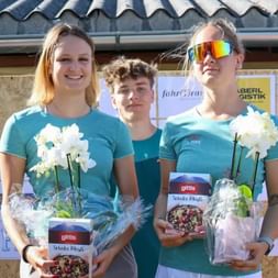 Three athletes Nici, Manuel and Lea in turquoise shirts holding white orchids and prizes on podium at Grabenseelauf 2025 event.