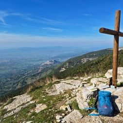 Holzkreuz auf felsigem Gipfel des Monte Subasio in Umbrien mit blauem Rucksack am Fuß. Panoramablick auf grüne Täler und Hügel unter klarem Himmel.