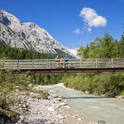 Zwei Wanderer stehen auf Holzbrücke über türkisfarbene Leutascher Ache mit dramatischen Kalksteinbergen und Wald im Hintergrund.