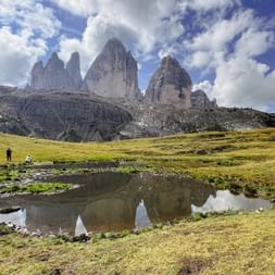 Drei Zinnen in den Dolomiten spiegeln sich in einem kleinen Teich, mit grünen Wiesen und zwei Wanderern unter dramatischem Wolkenhimmel.
