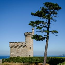 Steinerner Festungsturm mit Zinnen neben einer hohen Kiefer mit Blick auf den Atlantik bei Baiona, mit fernen Inseln am Horizont.
