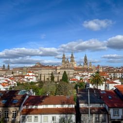 Panoramablick auf Santiago de Compostela mit den Türmen der Kathedrale über den roten Ziegeldächern unter blauem Himmel mit weißen Wolken.