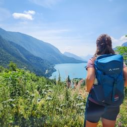 Female hiker with blue backpack viewing turquoise Lake Molveno surrounded by forested mountains under blue sky.
