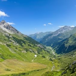 Panoramic view of a green alpine valley with winding paths, surrounded by rocky mountain peaks under a blue sky with white clouds.