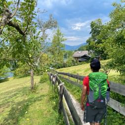 Wanderer mit grünem Rucksack auf eingezäuntem Weg durch Wiese im Salzkammergut, mit Blick auf See, Berge und traditionelles Haus.