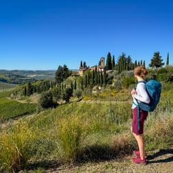 Female hiker with blue backpack standing on hillside path, viewing Tuscan villa with cypress trees and rolling green hills under clear blue sky.