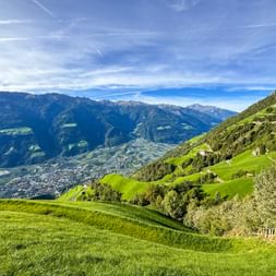 Panoramic view from Meraner Höhenweg showing green alpine meadows, scattered farmhouses, and mountain ranges under blue sky with valley below.