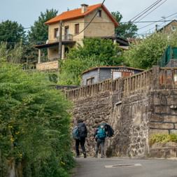 Zwei Wanderer mit Rucksäcken gehen auf einem gepflasterten Weg unter einer Steinbrücke in Arcade am Portugiesischen Jakobsweg.