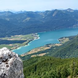 Aerial view of turquoise Wolfgangsee surrounded by green meadows and forested mountains. Rocky outcrop in foreground, Alpine peaks in distance.