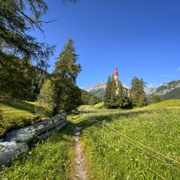Wanderweg entlang eines Gebirgsbachs im Obernberger Tal mit Kirche mit rotem Turm, umgeben von Wiesen und Alpengipfeln.