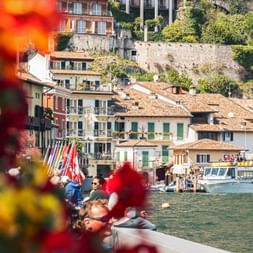 View of Limone sul Garda waterfront with colorful buildings, boats on Lake Garda, and people on the promenade framed by red flowers.